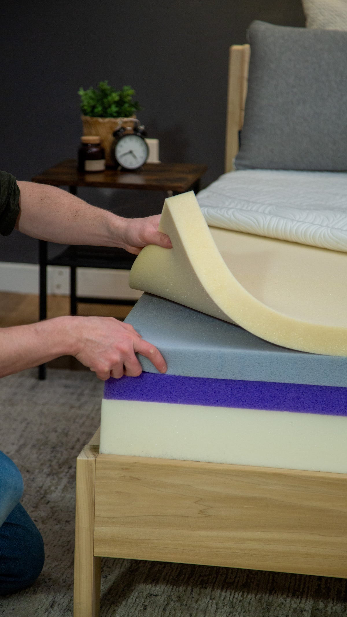 Person arranging foam layers on a wooden platform with a gray couch and plant in the background.