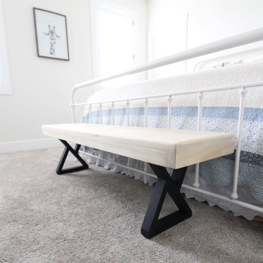 Wooden bench with black metal legs in front of a white bed with blue bedding.