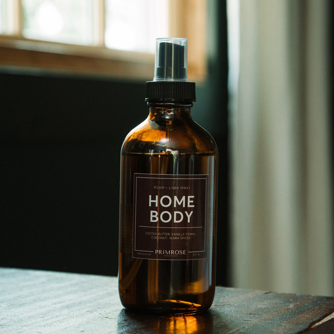 A bottle of Primrose Homebody Room Spray sitting on a dresser in front of a window