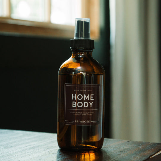 A bottle of Primrose Homebody Room Spray sitting on a dresser in front of a window