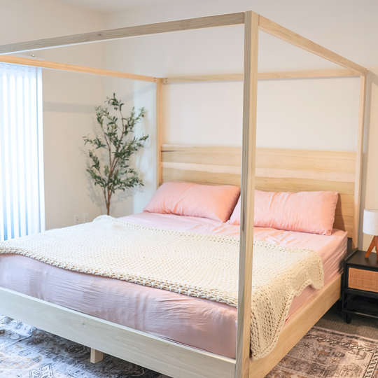 Wooden canopy bed with pink bedding in a room with a plant and nightstand.