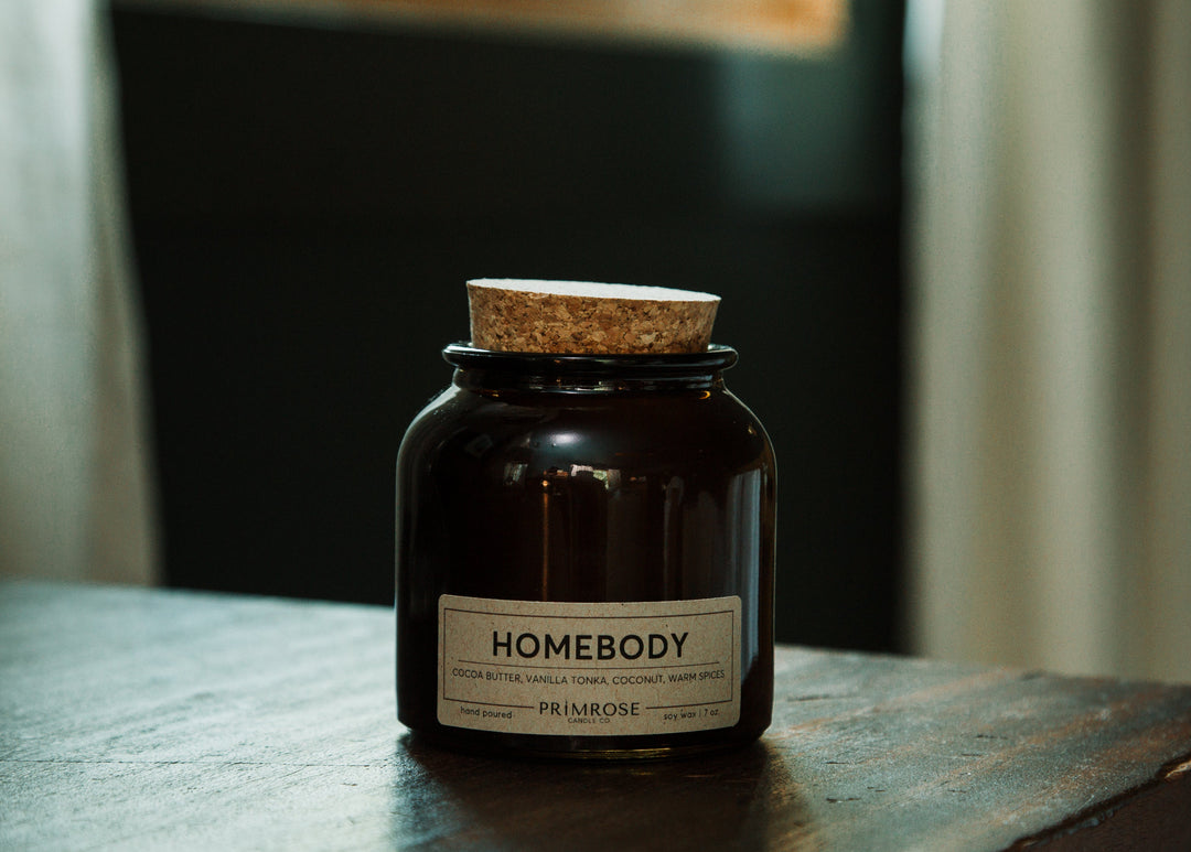 A dark amber glass jar candle with a cork lid and a white label reading 'HOMEBODY' and 'PRIMROSE', placed on a dark wooden table in a dimly lit interior setting.