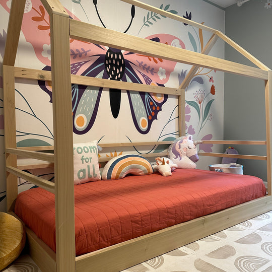 Wooden children's bed with red mattress against a wall with butterfly mural