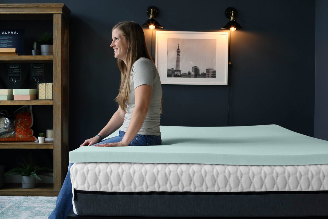 Woman sitting on a mattress in a bedroom with a bookshelf and framed picture in the background.
