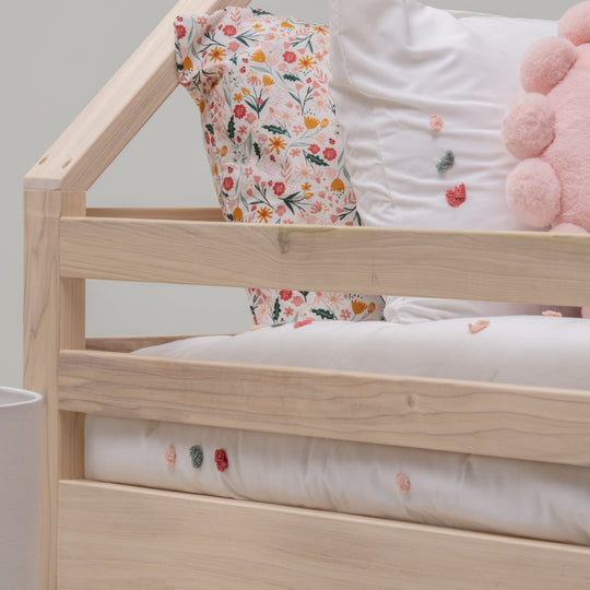 Wooden crib with floral-patterned mattress and pink plush toy against a plain wall.