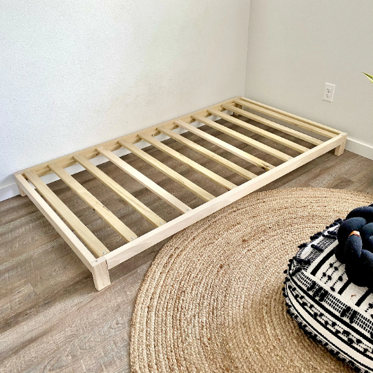 Wooden bed frame on a wooden floor with a round rug and black and white patterned pillow.