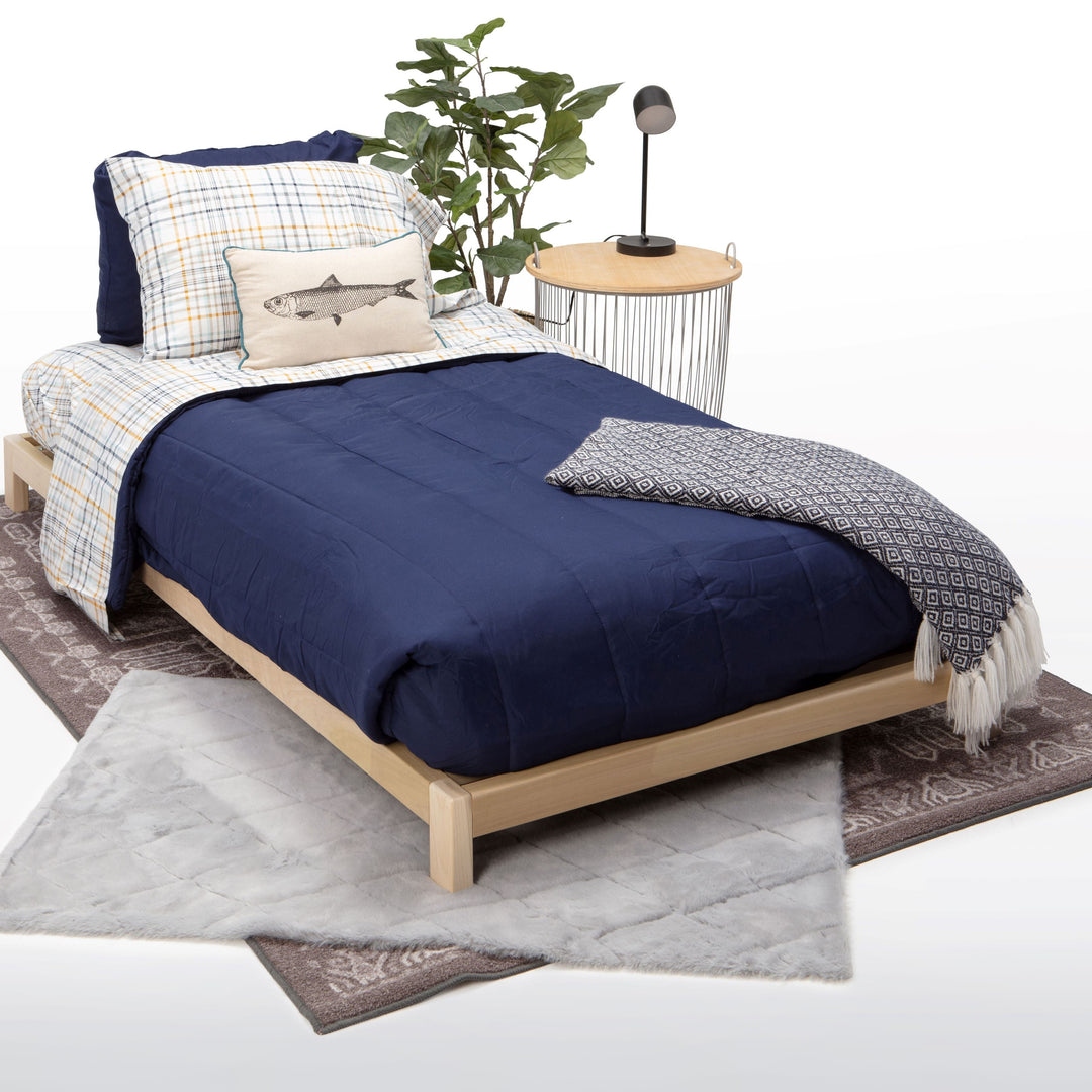 Bed with blue bedding and pillows, side table with plant, and rug on a white background