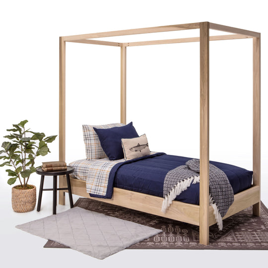 Wooden canopy bed with blue bedding and pillows, accompanied by a plant and small table on a white background.