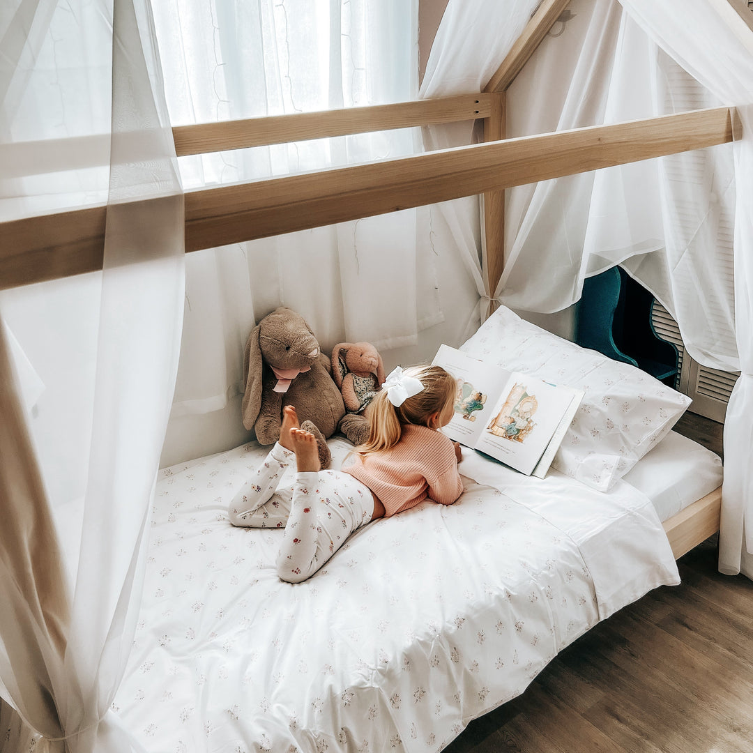 Child sitting on a bed with a wooden frame and white curtains, holding a teddy bear.